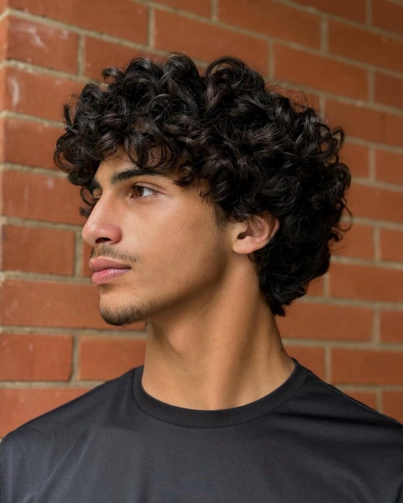 Young man with voluminous dark curly hair posing against a red brick wall.