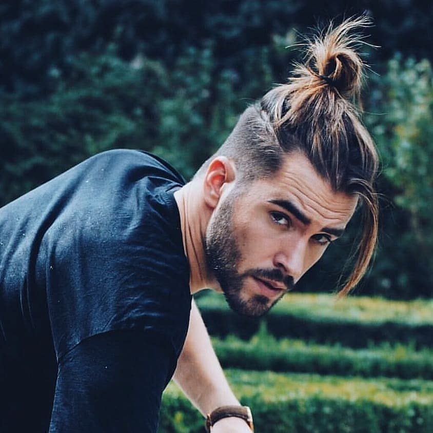 Man with a brown beard and a man-bun hairstyle posing outdoors in nature.
